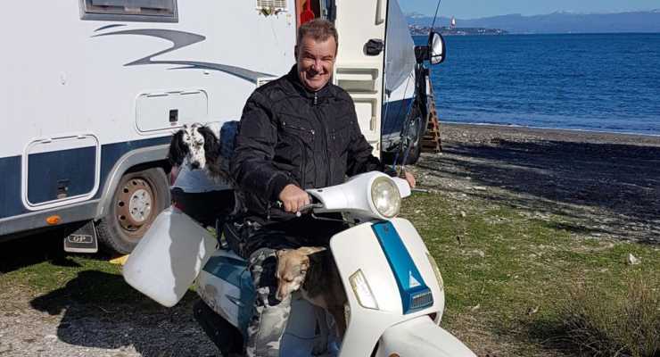 Man på vespa eller EU-moppe med hundar framför husbil och hav