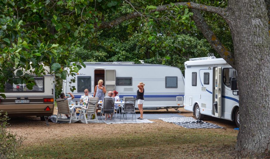 två husvagnar och en husbil på en campingplats, personer vid utemöbler framför dem